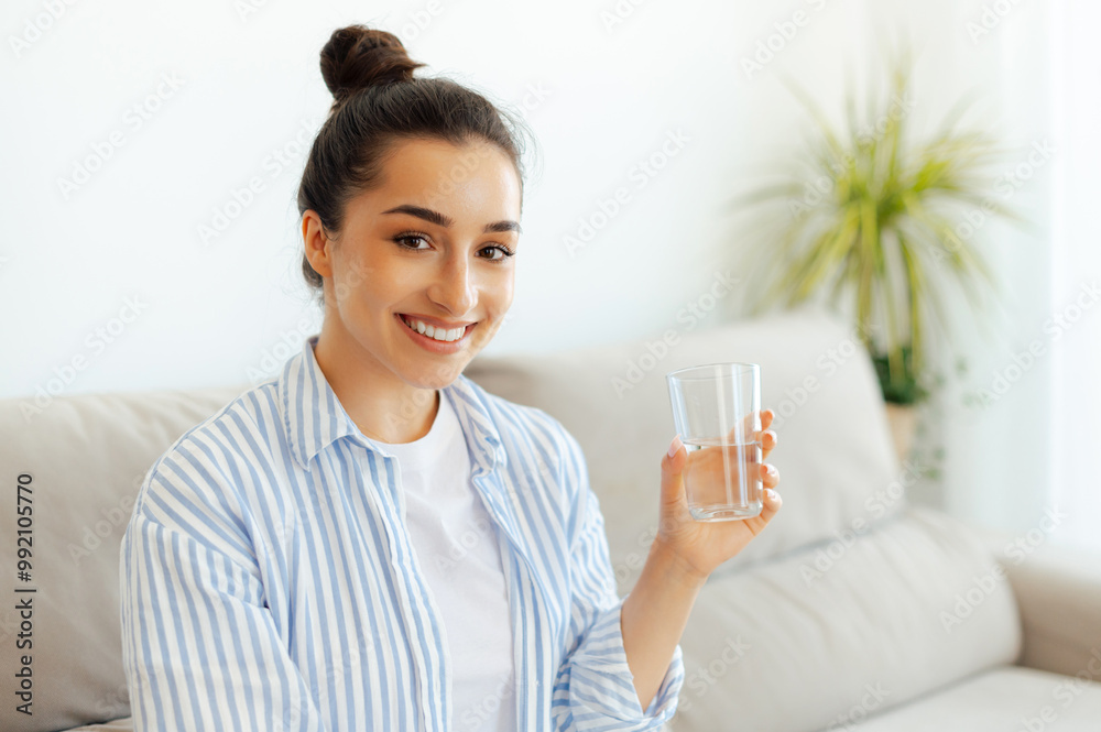 Healthy life style concept. Beautiful positive arabian or indian brunette woman in casual wear is holding a glass cup of water on the morning, while sits in a couch in a living room, smiles at camera Healthy life style concept. Beautiful positive arabian or indian brunette woman in casual wear is holding a glass cup of water on the morning, while sits in a couch in a living room, smiles at camera