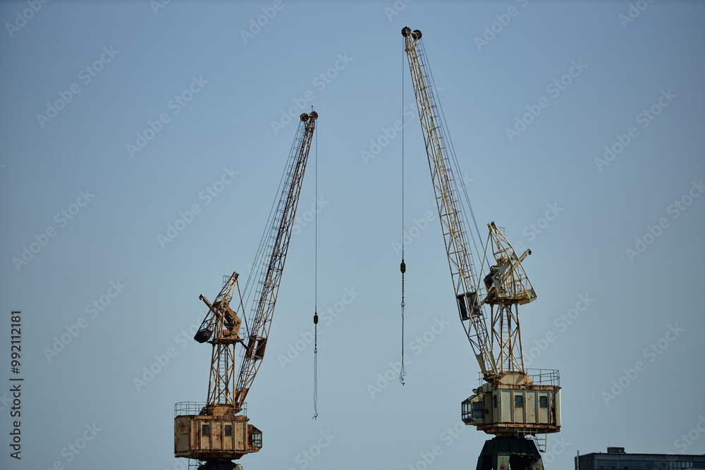 Dual cranes standing tall against clear sky forming prominent vertical lines symbolizing active construction site creating industrial backdrop emphasizing urban development Dual cranes standing tall against clear sky forming prominent vertical lines symbolizing active construction site creating industrial backdrop emphasizing urban development
