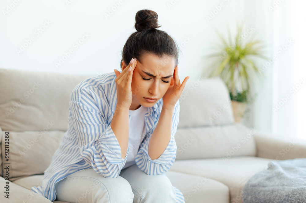 Stress, headache, illness. Upset mixed race woman sitting on a sofa in living room with eyes closed, suffering headache, vertigo, migraine, having cramp in a templess, need treatment and rest Stress, headache, illness. Upset mixed race woman sitting on a sofa in living room with eyes closed, suffering headache, vertigo, migraine, having cramp in a templess, need treatment and rest