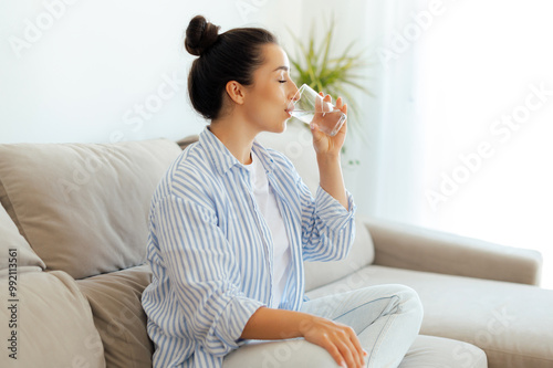 Beautiful happy calm arabian or indian brunette woman in casual wear is drinking a glass cup of clean water in the morning, while sits on sofa in living room at home, leads healthy life style concept