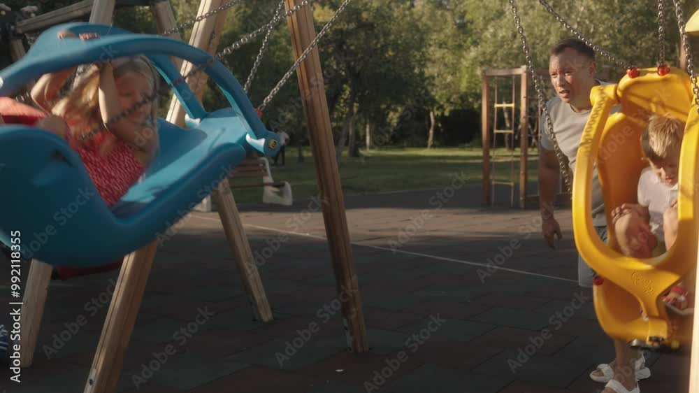 A father pushes his two young children on swings at the playground. They are all smiles and laughter on a sunny summer day. Perfect for concepts about family, childhood, and summer fun. A father pushes his two young children on swings at the playground. They are all smiles and laughter on a sunny summer day. Perfect for concepts about family, childhood, and summer fun.