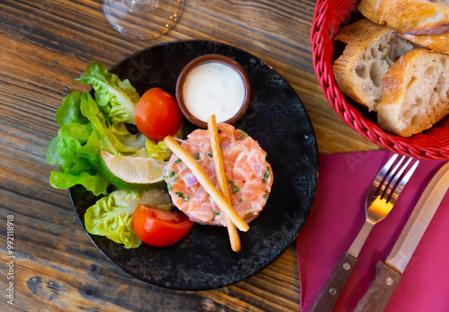 Raw salmon tartare seasoned with cream chive sauce served with slices of fresh tomatoes, green lettuce, lime and crispy bread sticks. French fish appetizer photo