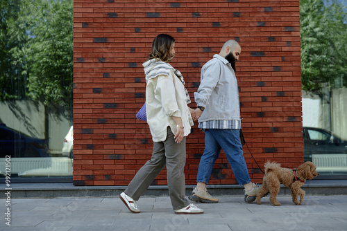 Canvas-taulu Two individuals walking on city sidewalk with small dog walking alongside