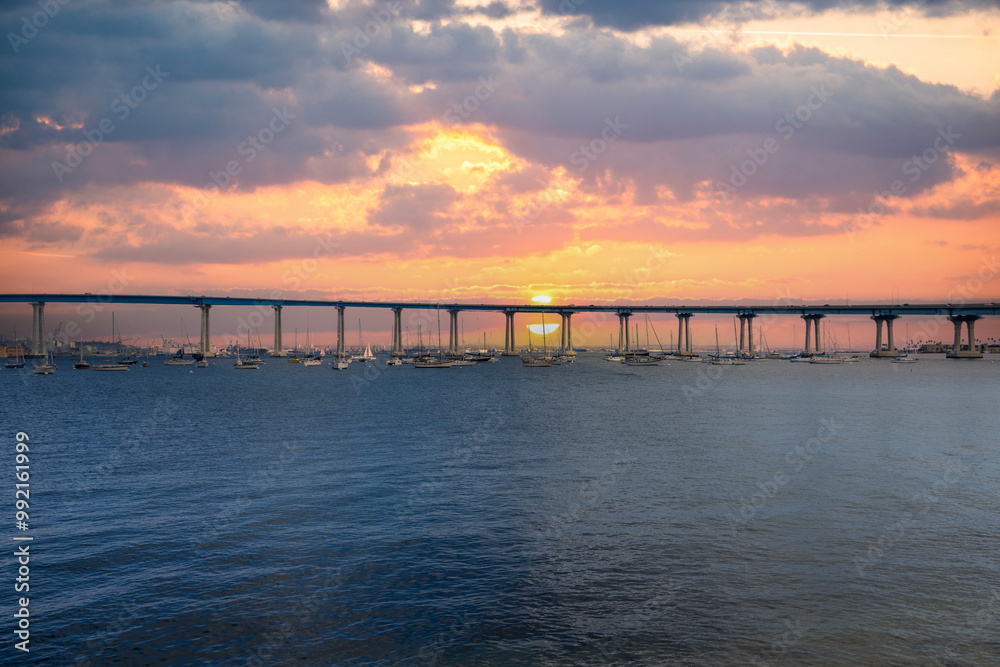 Fototapeta premium San Diego bay and the Coronado Bridge with ships sailing on blue ocean water, office buildings and skyscrapers along the coast at sunset at Coronado Tidelands Park in Coronado California