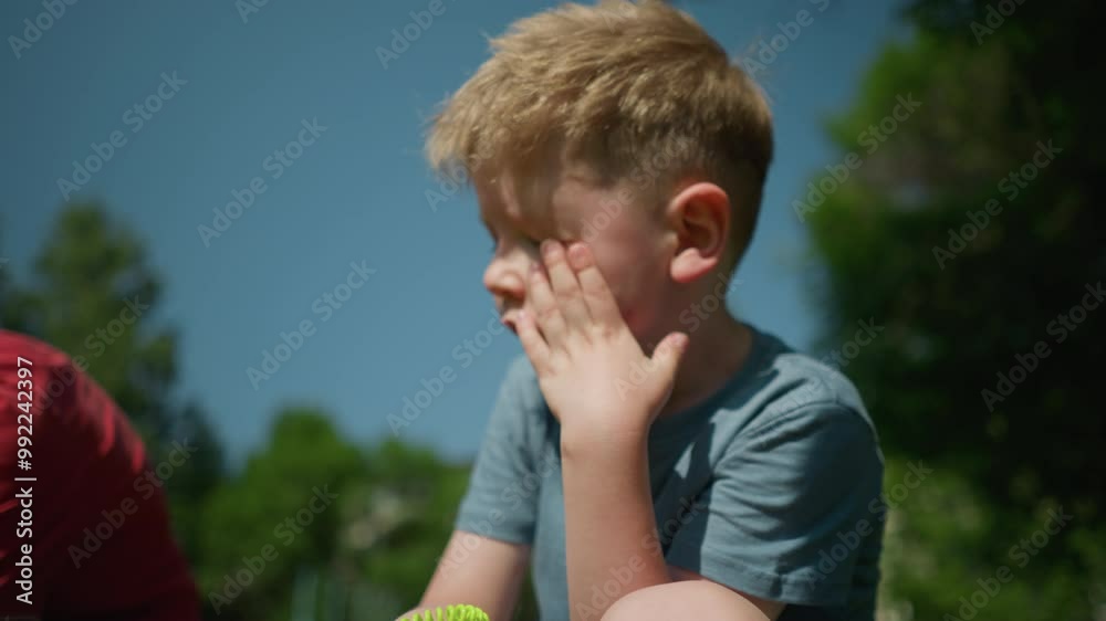 A little boy wearing a blue shirt rubs his face with his left hand, sitting in a grassy field with a partial view of someone close by him A little boy wearing a blue shirt rubs his face with his left hand, sitting in a grassy field with a partial view of someone close by him