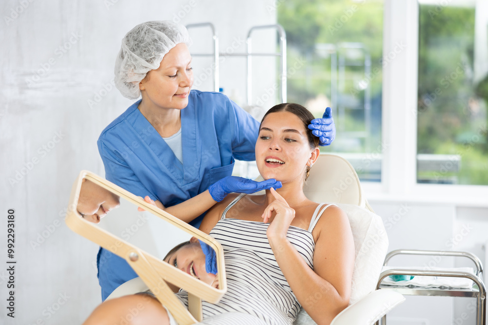 Fototapeta premium Adult female doctor cosmetologist analyzes face of young female patient in medical office