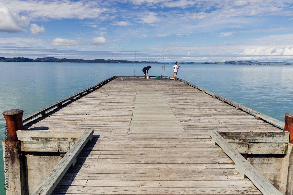 Obraz premium People fishing off a pier over a calm ocean beach in Maraetai, Auckland, New Zealand.