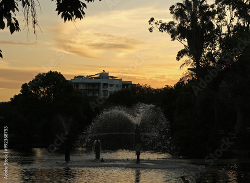 Sunset scene with fountains spraying water in a creek surrounded by trees and a modern building in the background, Redcliffe, Queensland, Australia photo