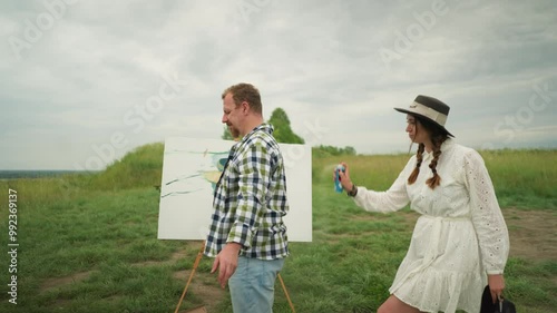 A woman in a stylish hat and white dress sprays mosquito repellent on a man s head in a grass field. The man, in a checkered shirt, stands calmly as she sprays. A painting board and chair are visible photo