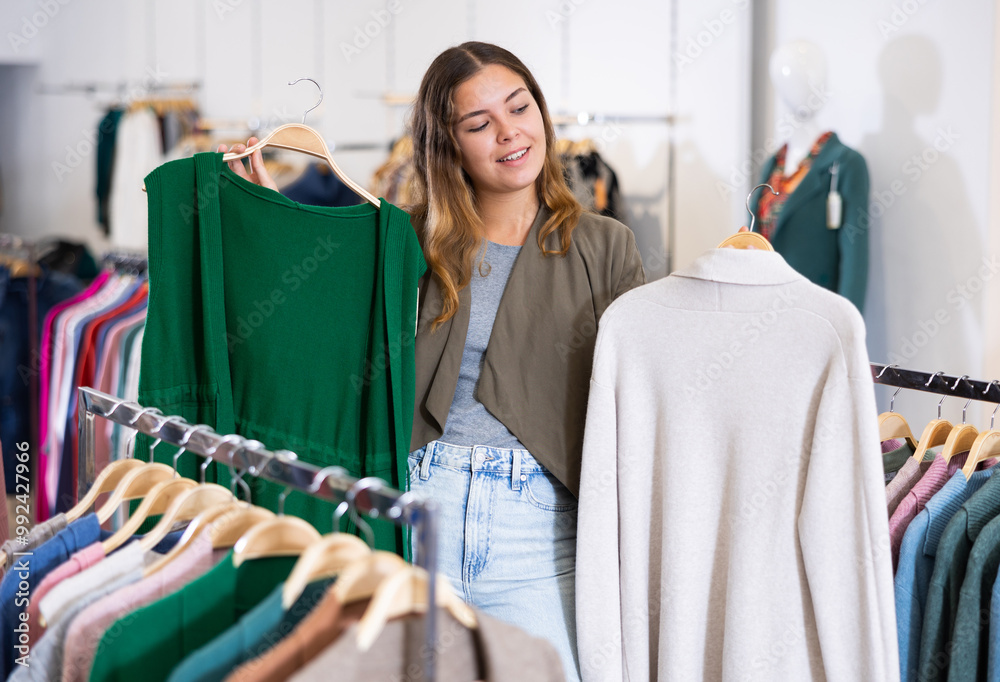 Fototapeta premium Curious young female choosing between green cardigan without sleeves and white sweater in a clothing store