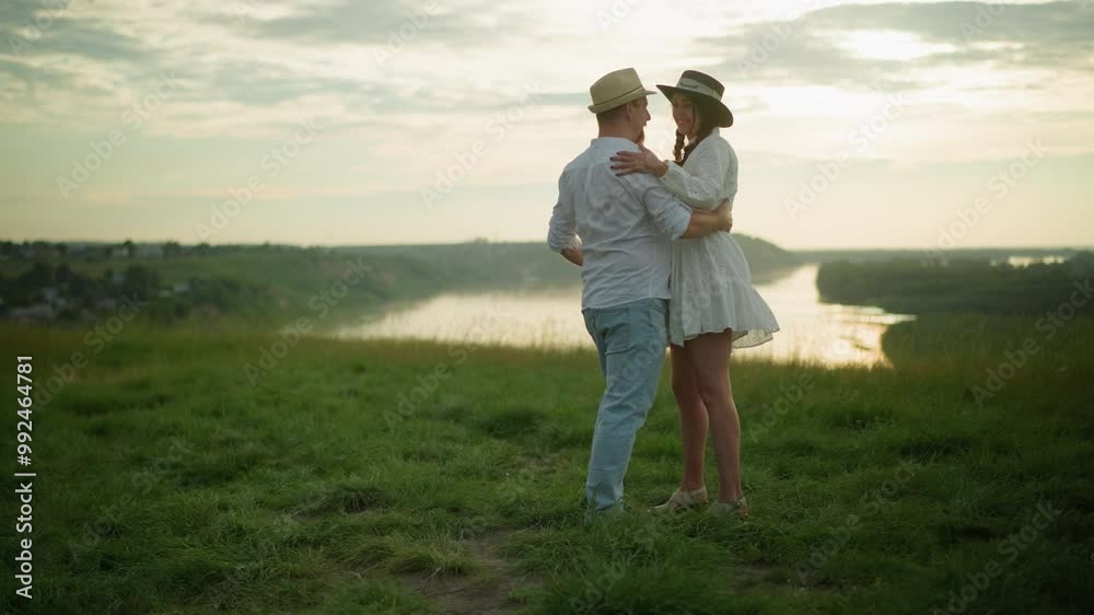 Two happy people hold each other while dancing on a grassy hill beside a tranquil lake. The man is dressed in a white shirt, hat, and jeans, while the woman wears a white dress and black hat Two happy people hold each other while dancing on a grassy hill beside a tranquil lake. The man is dressed in a white shirt, hat, and jeans, while the woman wears a white dress and black hat