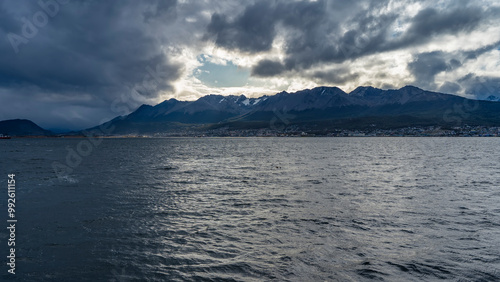 A picturesque snow-covered mountain range of the Andes against the sky and dark clouds. The city buildings of Ushuaia are at the foot. In the foreground is the surface of the ocean. The Beagle Channel