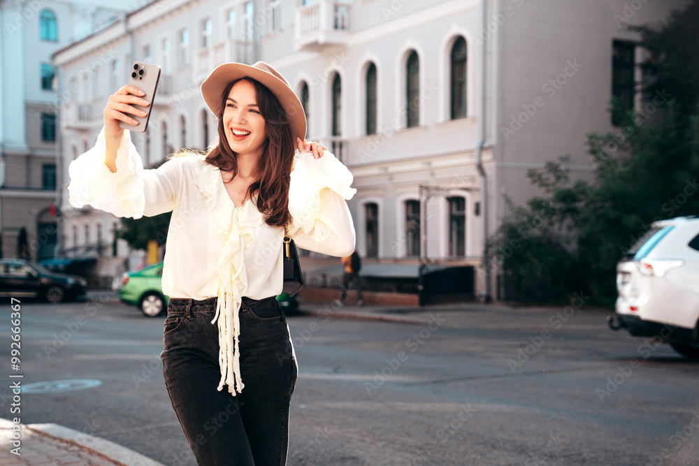 Young beautiful smiling hipster woman in trendy summer white blouse. Carefree woman posing in the street in sunny day. Positive model outdoors. Takes Pov selfie, makes photo holding camera in hand Young beautiful smiling hipster woman in trendy summer white blouse. Carefree woman posing in the street in sunny day. Positive model outdoors. Takes Pov selfie, makes photo holding camera in hand