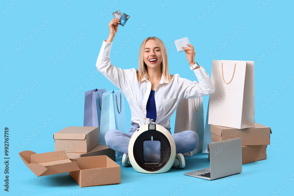 Young woman with laptop, shopping bags, wallet, credit cards and new coffee machine sitting on blue background