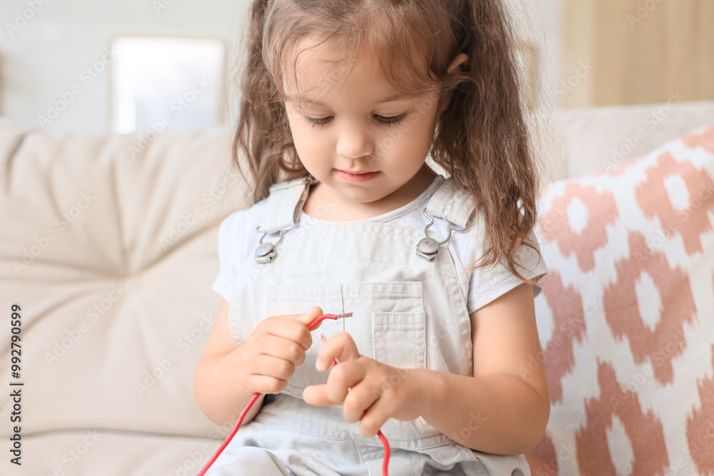 Cute little girl with bare wire sitting on sofa at home, closeup. Child in danger Cute little girl with bare wire sitting on sofa at home, closeup. Child in danger