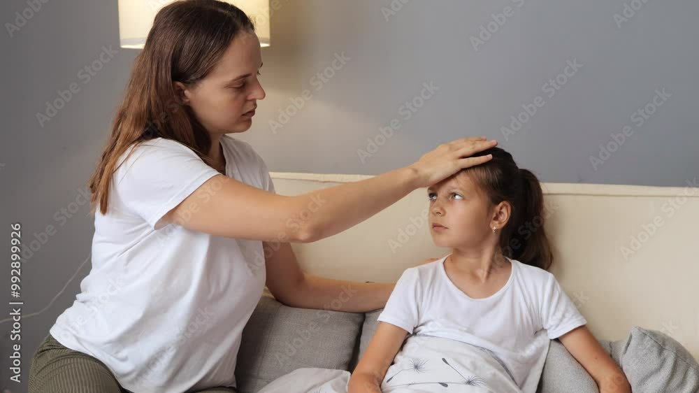 Child girl having fever mom taking care of child sitting together on sofa at their house frightened woman touching her little daughter's forehead to check temperature Child girl having fever mom taking care of child sitting together on sofa at their house frightened woman touching her little daughter's forehead to check temperature