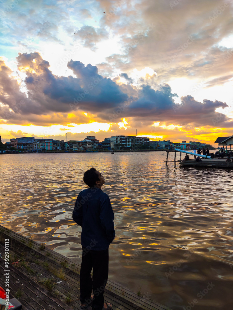 A person stands by the water, watching a vibrant sunset with colorful clouds reflecting on the calm surface, while city buildings create a peaceful, scenic backdrop.