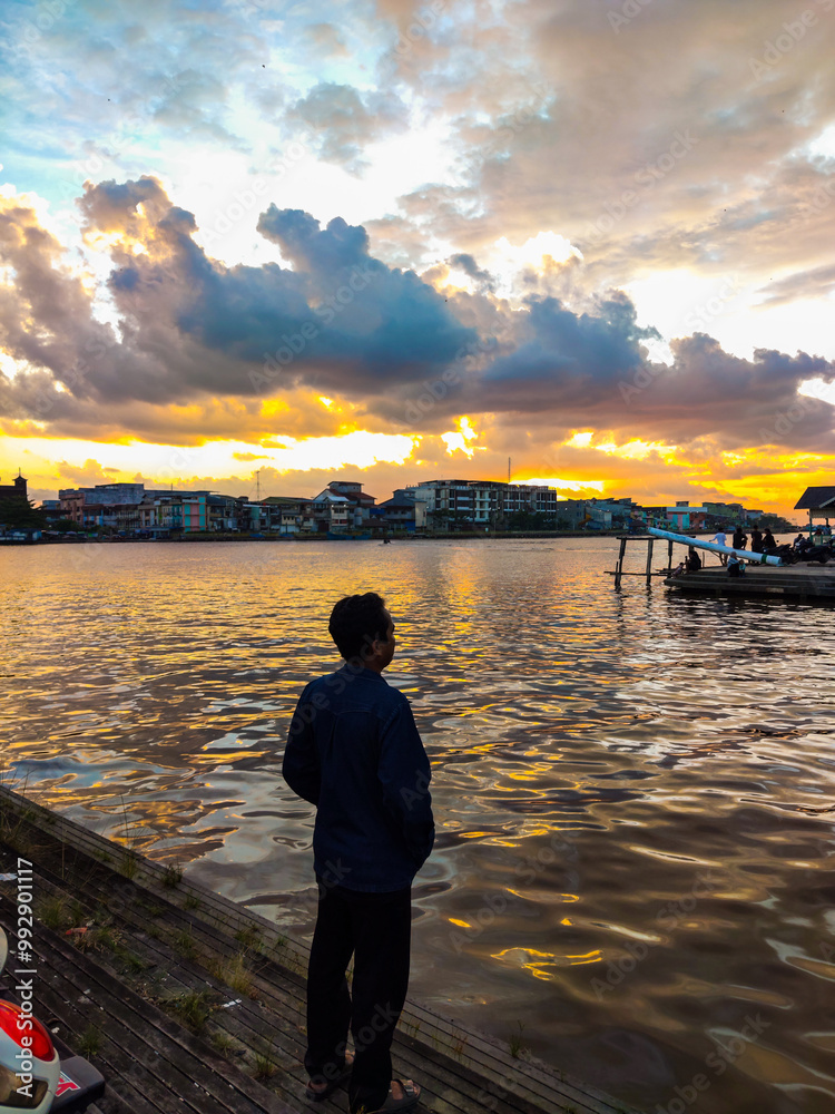 A person stands by the water, watching a vibrant sunset with colorful clouds reflecting on the calm surface, while city buildings create a peaceful, scenic backdrop.