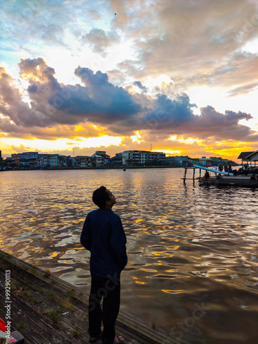 Obraz na płótnie A person stands by the water, watching a vibrant sunset with colorful clouds reflecting on the calm surface, while city buildings create a peaceful, scenic backdrop. w salonie