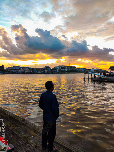 Obraz na płótnie A person stands by the water, watching a vibrant sunset with colorful clouds reflecting on the calm surface, while city buildings create a peaceful, scenic backdrop. w salonie