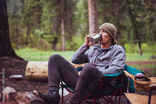 Man sitting in a camp chair around a fire drinking from a can