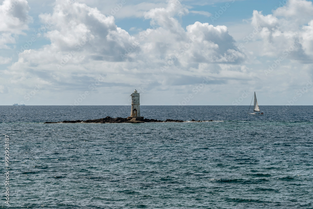 Naklejka premium A sailboat passes near the Mangiabarche lighthouse, Sant'Antioco island, Italy
