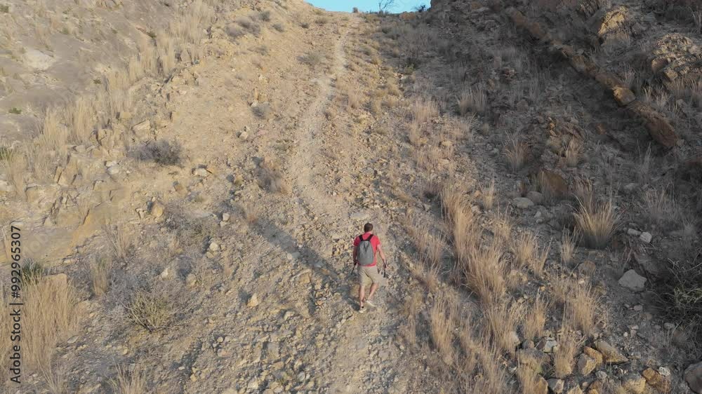A man in a red shirt and shorts, carrying trekking poles and a backpack, climbs a rocky mountain trail in the morning sun. A solitary hike for calm and fitness.