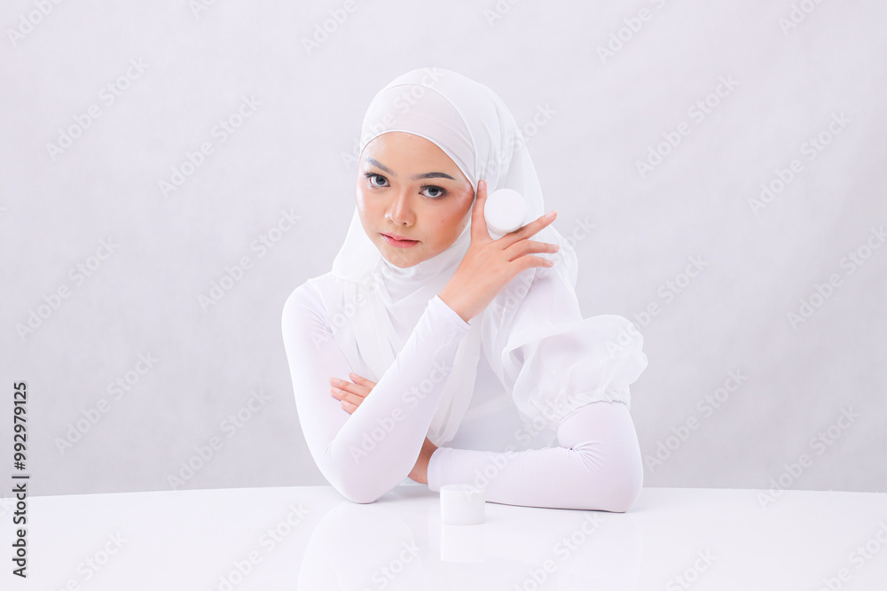 portrait of beautiful young woman in hijab elegant looking at camera hand holding cosmetic cream for daily facial care routine on white background. advertising, health, medical and fashion concept portrait of beautiful young woman in hijab elegant looking at camera hand holding cosmetic cream for daily facial care routine on white background. advertising, health, medical and fashion concept