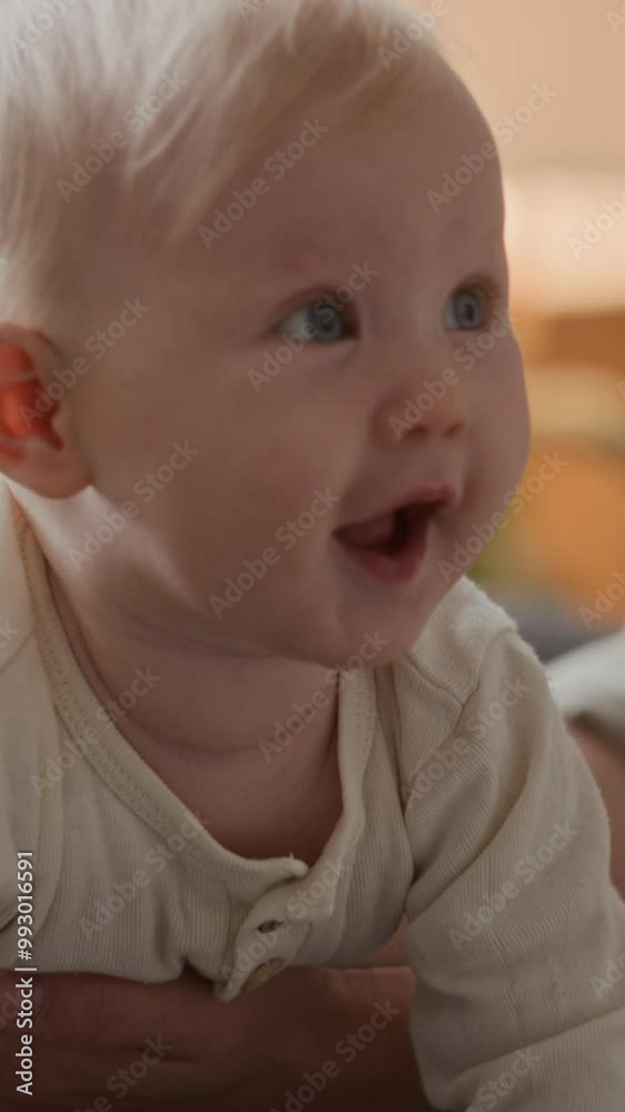 Vertical close-up of adorable curious Caucasian baby boy or girl smiling while sitting on couch with unrecognizable mother supporting and preventing child from falling Vertical close-up of adorable curious Caucasian baby boy or girl smiling while sitting on couch with unrecognizable mother supporting and preventing child from falling