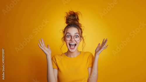 Photo of a funny excited young woman with an open mouth and raised hands wearing glasses against a yellow background photo