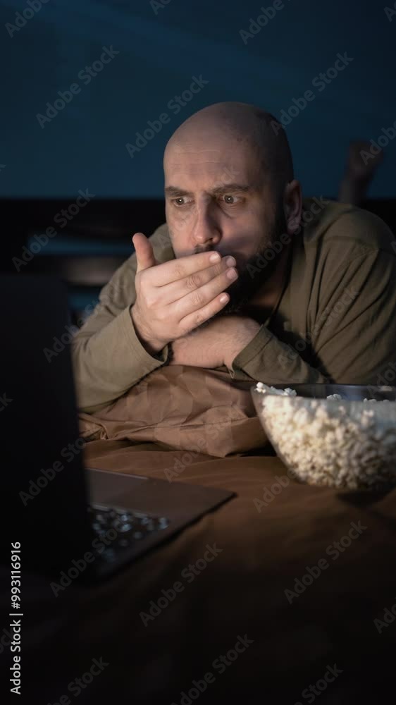 Man looks at laptop screen feels angry due team loss, lying in bed at night eating popcorn resting in bedroom Man looks at laptop screen feels angry due team loss, lying in bed at night eating popcorn resting in bedroom