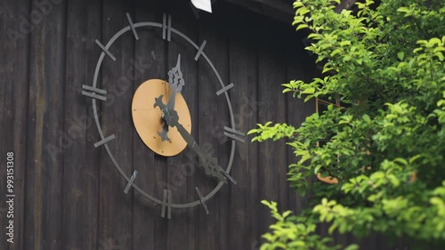 A large outdoor clock with decorative hands is mounted on a wooden wall, surrounded by greenery. photo