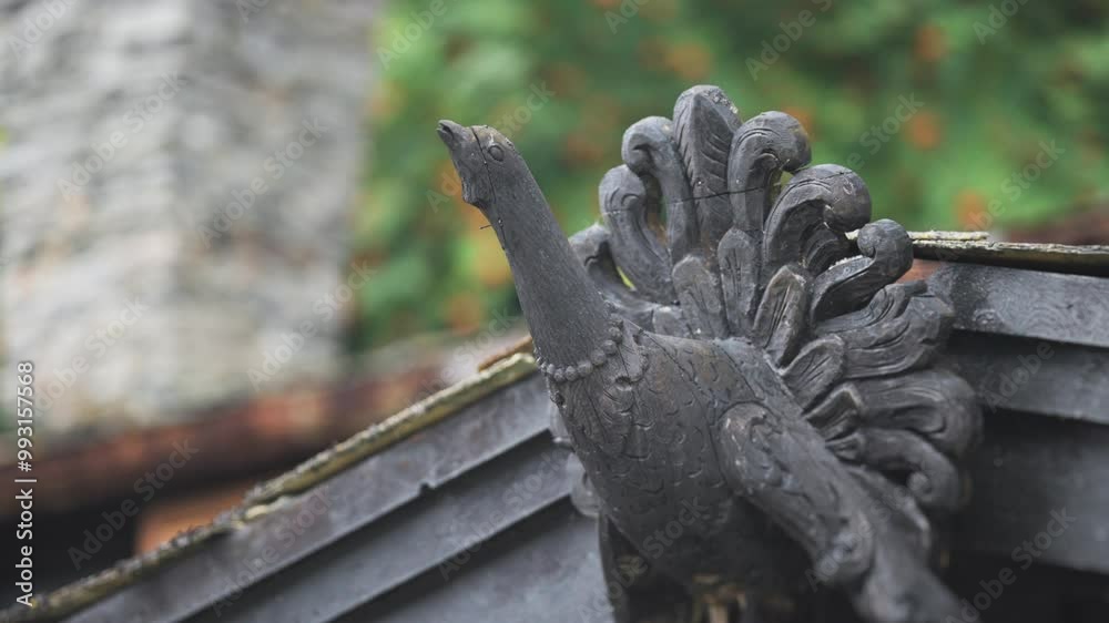 An intricately carved peacock decorates the top of a structure, its tail fanned out in a natural outdoor setting. An intricately carved peacock decorates the top of a structure, its tail fanned out in a natural outdoor setting.