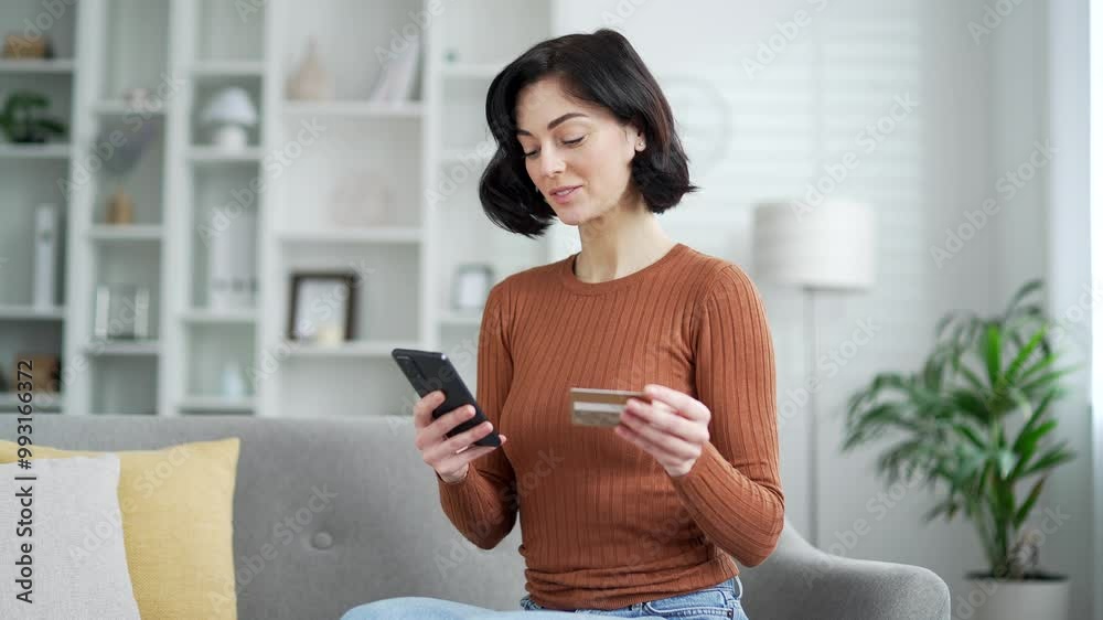 Happy young adult woman enjoying online shopping experience with phone and credit card sitting on sofa in living room at home. Smiling female happily makes a purchase and looks into the camera Happy young adult woman enjoying online shopping experience with phone and credit card sitting on sofa in living room at home. Smiling female happily makes a purchase and looks into the camera