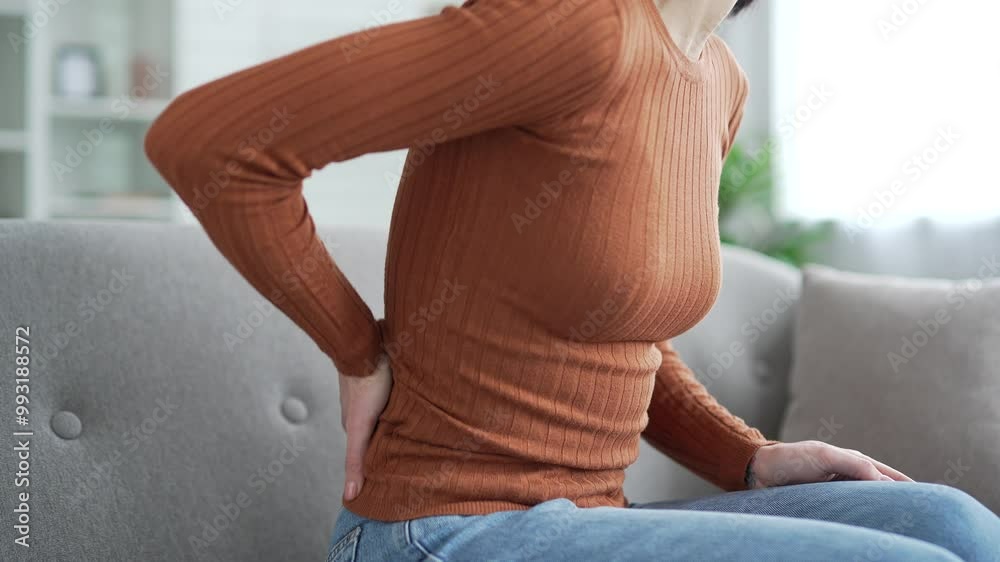 Close up. Female hand holding massaging lower back. Tired young adult woman suffering from back pain while sitting on sofa in living room at home. Upset lady shows discomfort due to poor posture