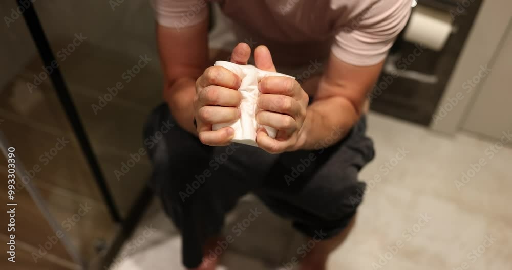 Male person grips toilet paper tightly sitting on toilet bowl in apartment bathroom. Concept of daily routine in modern premise slow motion