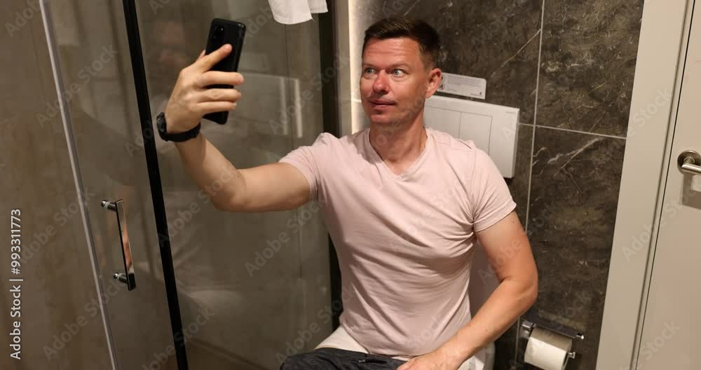 Male person takes funny selfie sitting on toilet bowl in light bathroom. Man poses for photo on mobile phone in premise of apartment slow motion