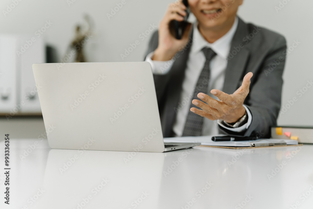 Confident Businessman on a Call:  A focused businessman in a sharp suit sits at his desk, actively engaged in a phone call while a laptop rests open before him.