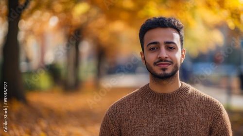 A young man stands confidently in a warm sweater, smiling against a backdrop of vibrant autumn foliage. Sunlight filters through the trees, creating a picturesque fall atmosphere.