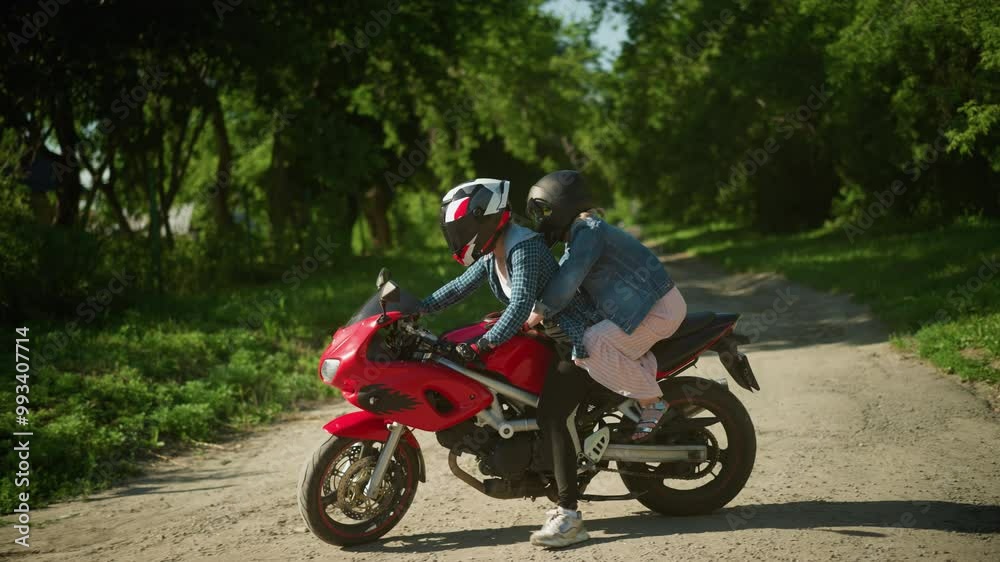 Two sisters ride a red power bike, both wearing helmets, as they drive into a paved walking path surrounded by trees, a blur view of buildings can be seen in the background Two sisters ride a red power bike, both wearing helmets, as they drive into a paved walking path surrounded by trees, a blur view of buildings can be seen in the background