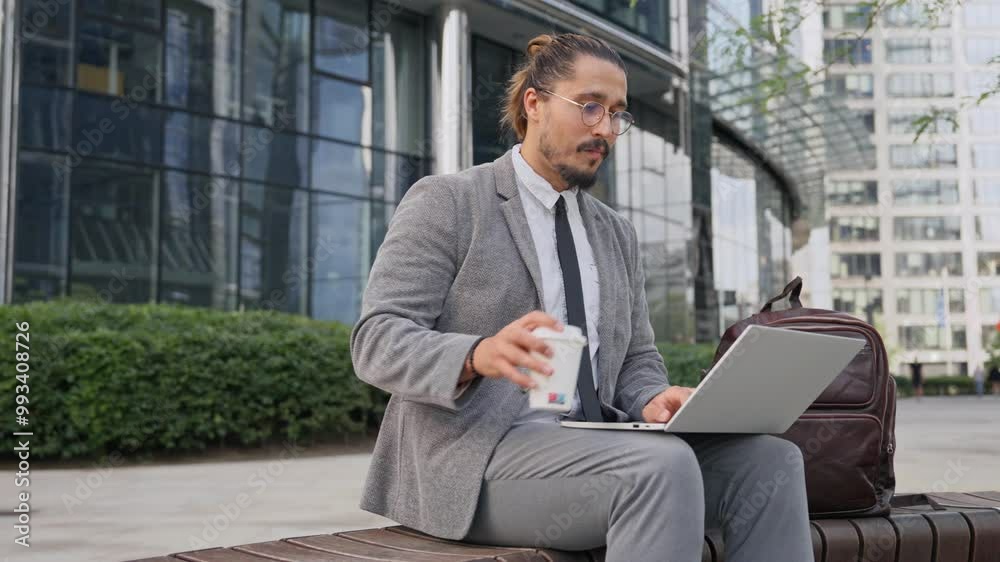 Side view of Caucasian man sitting on bench in middle of big building block. Using laptop and taking sip of hot drink. Working in fresh air. Freelancing after work. Searching for information online. Side view of Caucasian man sitting on bench in middle of big building block. Using laptop and taking sip of hot drink. Working in fresh air. Freelancing after work. Searching for information online.