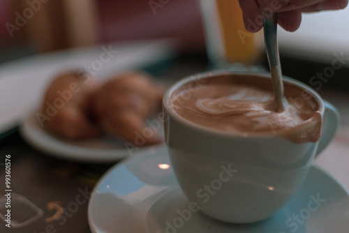 Una mano agita suavemente una taza de café con leche, revelando un diseño floral en la espuma. Un delicioso capuchino en un ambiente acogedor, perfecto para comenzar el día