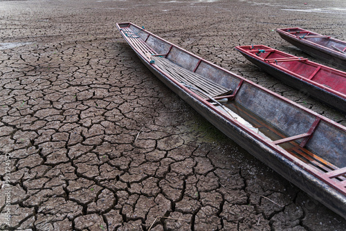 The ground is cracked and parched, with a network of deep fissures. Several traditional wooden boats are beached on the dried-up water body, highlighting the severity of the drought.