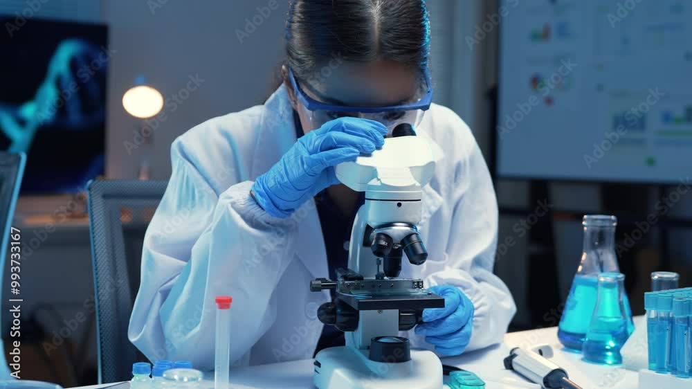 Scientist wearing a lab coat and safety glasses adjusts a microscope in a well equipped laboratory setting Scientist wearing a lab coat and safety glasses adjusts a microscope in a well equipped laboratory setting