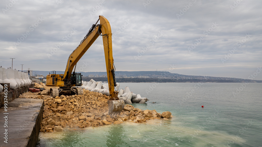 Fototapeta premium Waterfront construction site with excavator. Excavator working on a breakwater construction