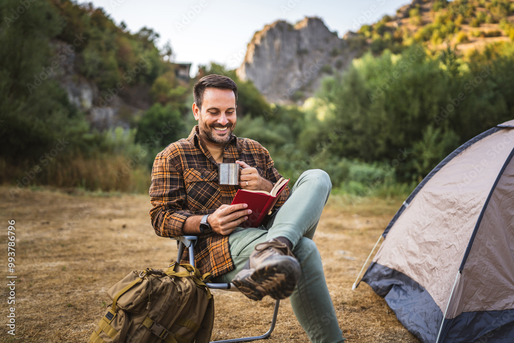 Fototapeta premium Adult caucasian man sit in front tent and read book and drink hot drink