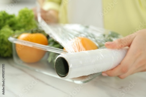 Woman putting plastic food wrap over glass container with fresh vegetables at white marble table, closeup
