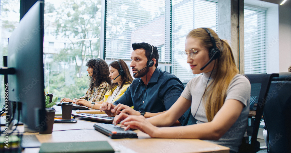 Fototapeta premium Hardworking dispatchers or consultants actively talking with clients on headsets. Attractive leader walking in background and controlling work process. People using computers at work. Support center.