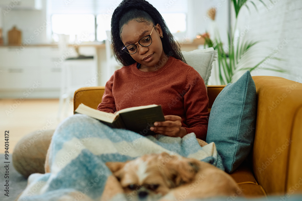 Young black woman reading book while relaxing with her dog at home. Young black woman reading book while relaxing with her dog at home.