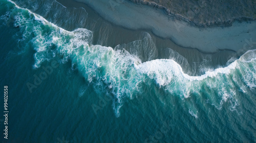 An overhead shot of the white foam of the waves crashing onto a sandy beach, with a clear separation between the deep ocean waters and the shallow waters offshore.